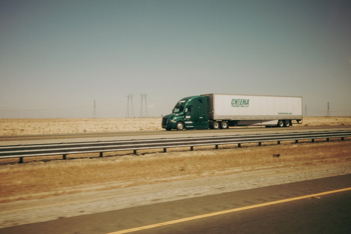 Green semi-truck on highway with dry landscape and wind turbines.
