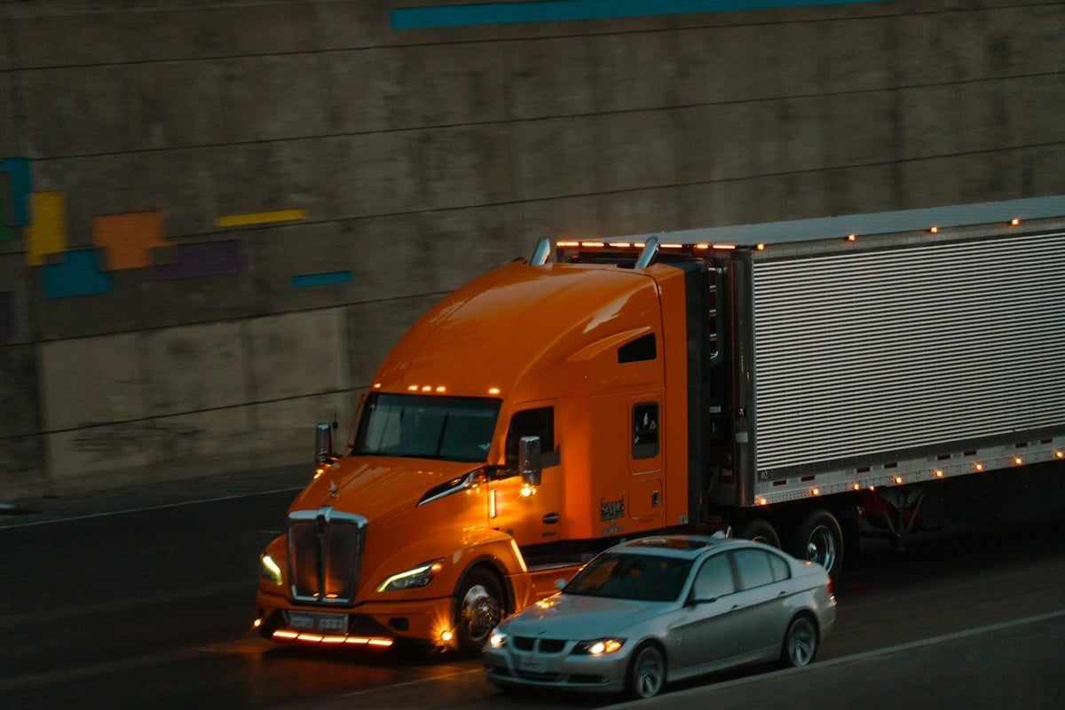 An orange semi-truck and a silver sedan driving on a highway at night.